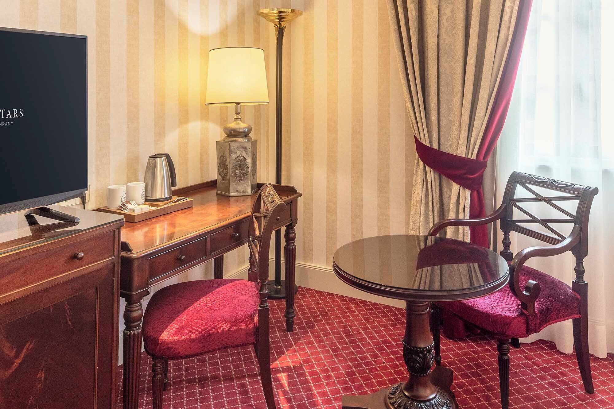 A hotel room with a flat-screen TV, a wooden desk with a lamp, a chair, and a small table.