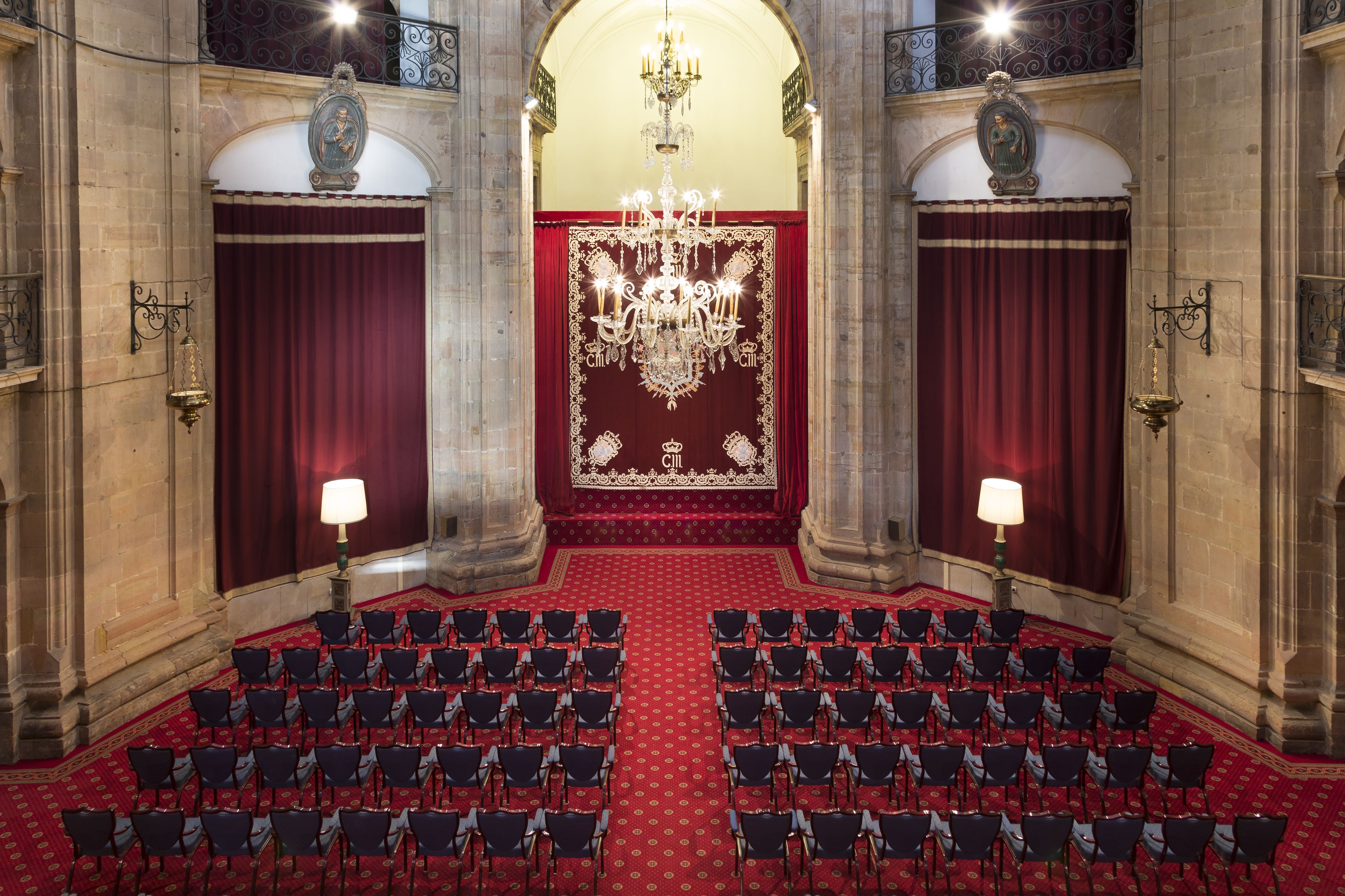 Un salón amplio con filas de sillas mirando hacia un escenario con alfombra roja y decoraciones elaboradas.