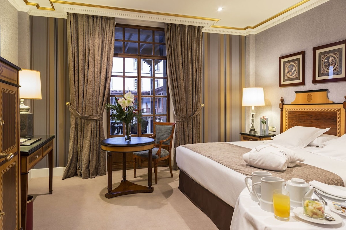 A hotel room with a bed, bedside table, chair, window with curtains, and a view of buildings outside.