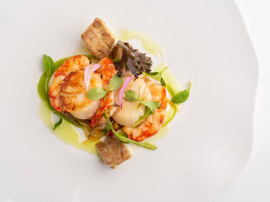 Un plato blanco con un guiso de mariscos que incluye camarones, vieiras y salsa verde.