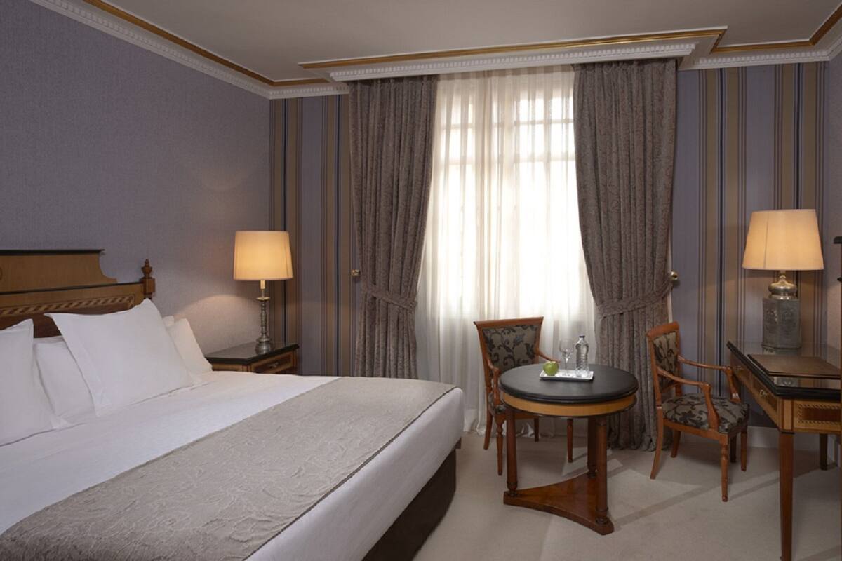 Ein Hotelzimmer mit einem Bett, zwei Nachttischen mit Lampen, einem Fernseher, einem kleinen runden Tisch mit Stühlen und einem großen Fenster mit Vorhängen.