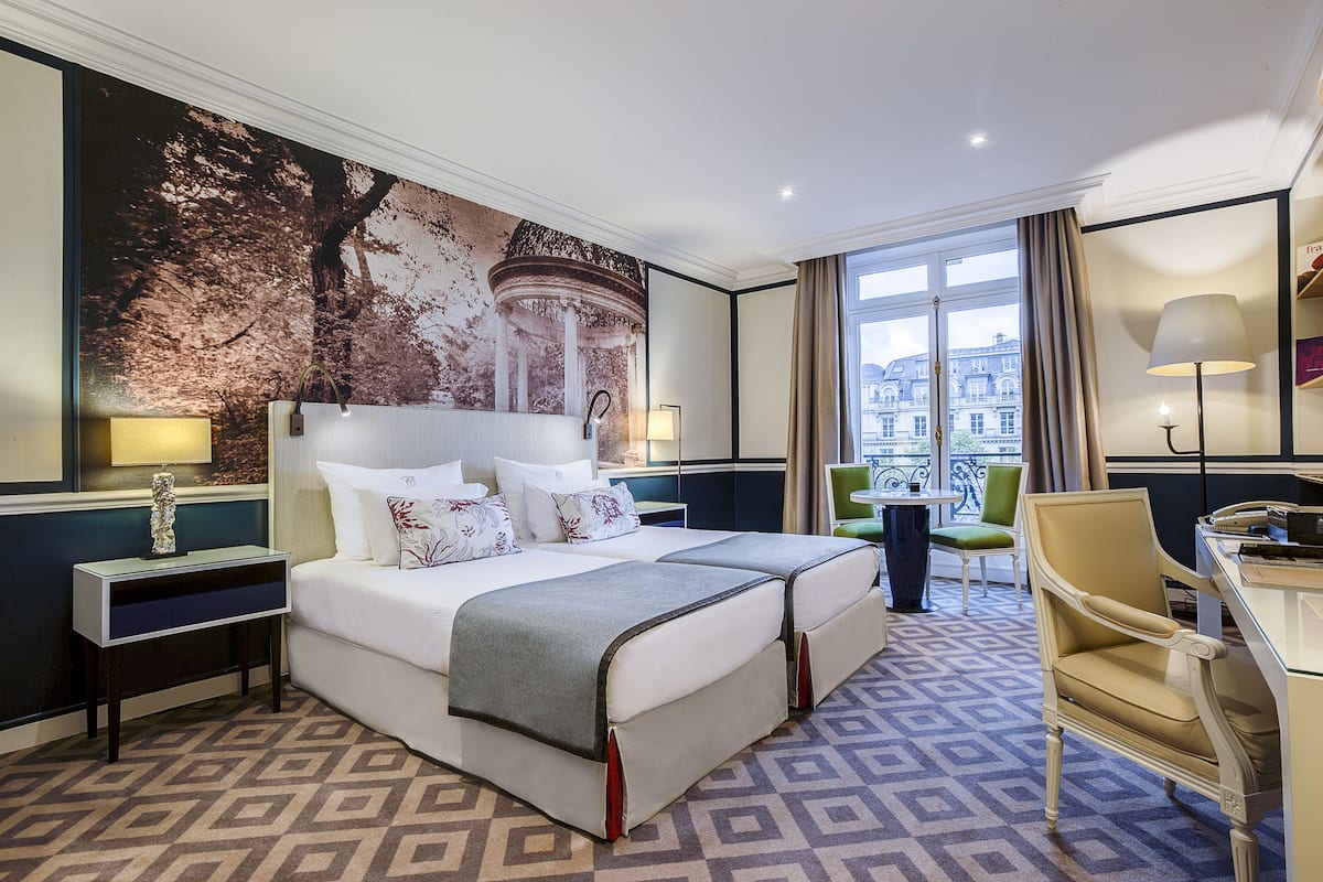 A hotel room with a large bed, two bedside tables, a desk with a chair, and a view of a building through the window.