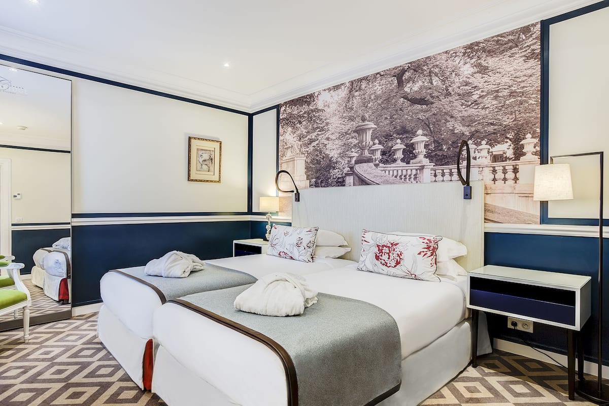 A hotel room with two beds, a large wall mural, a mirror, and a bedside table with a lamp.