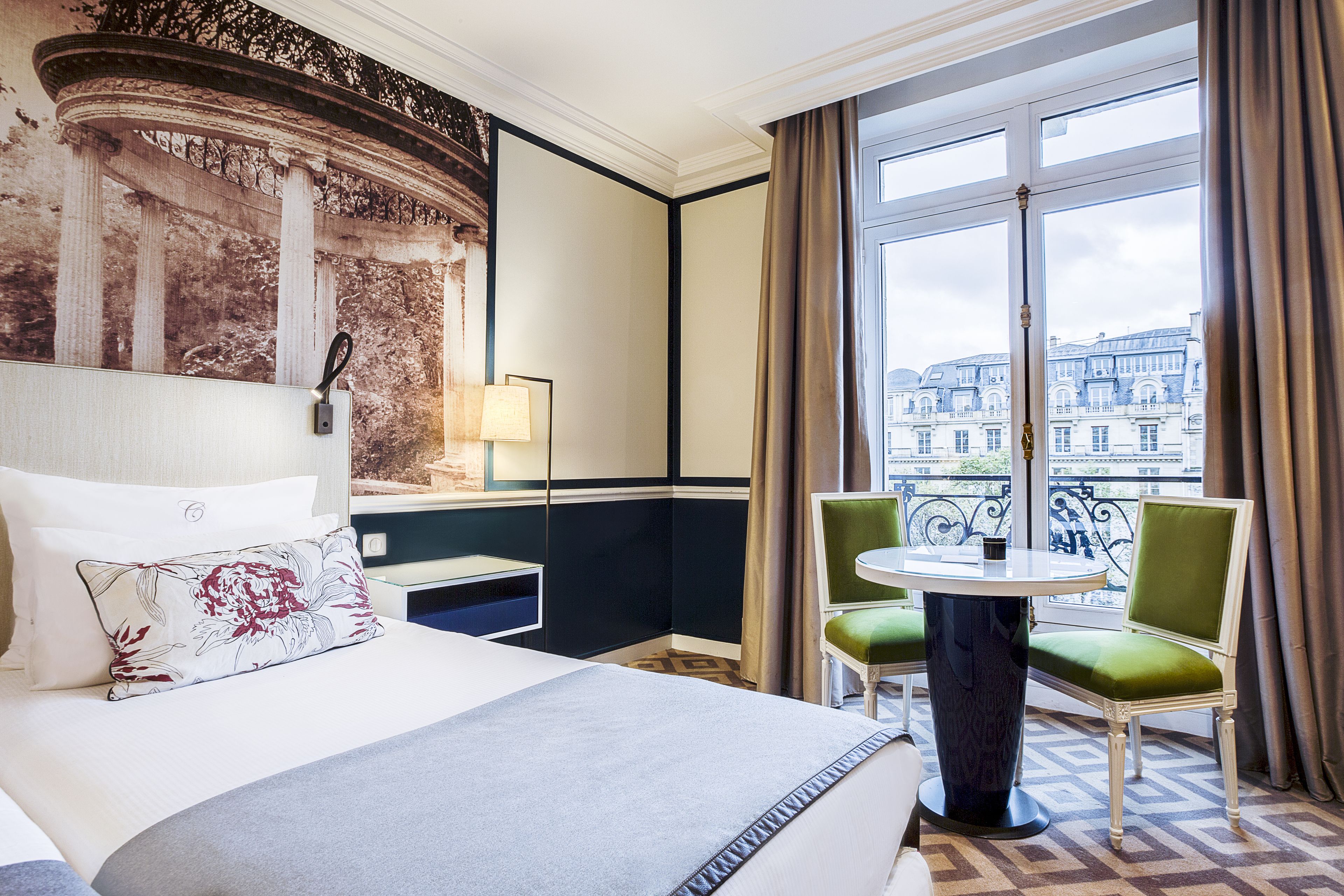 A hotel room with a large bed, a small table, two chairs, a view of a cityscape through a large window, and a decorative wall panel.