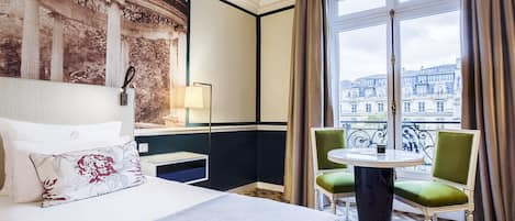 A hotel room with a large bed, a small table, two chairs, a view of a cityscape through a large window, and a decorative wall panel.