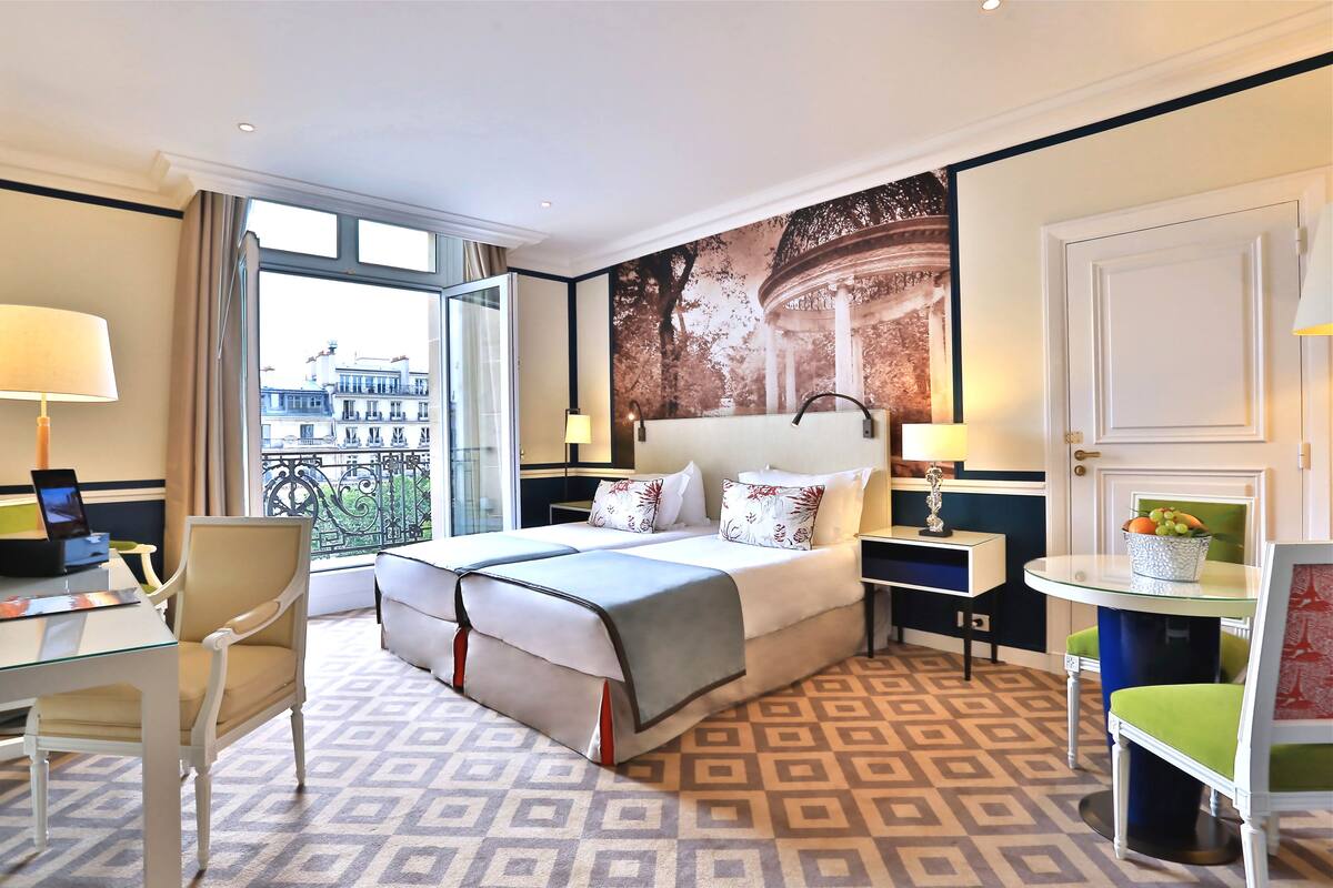 A hotel room with a large bed, a desk, a chair, a small table with a lamp, and a view of buildings through the window.