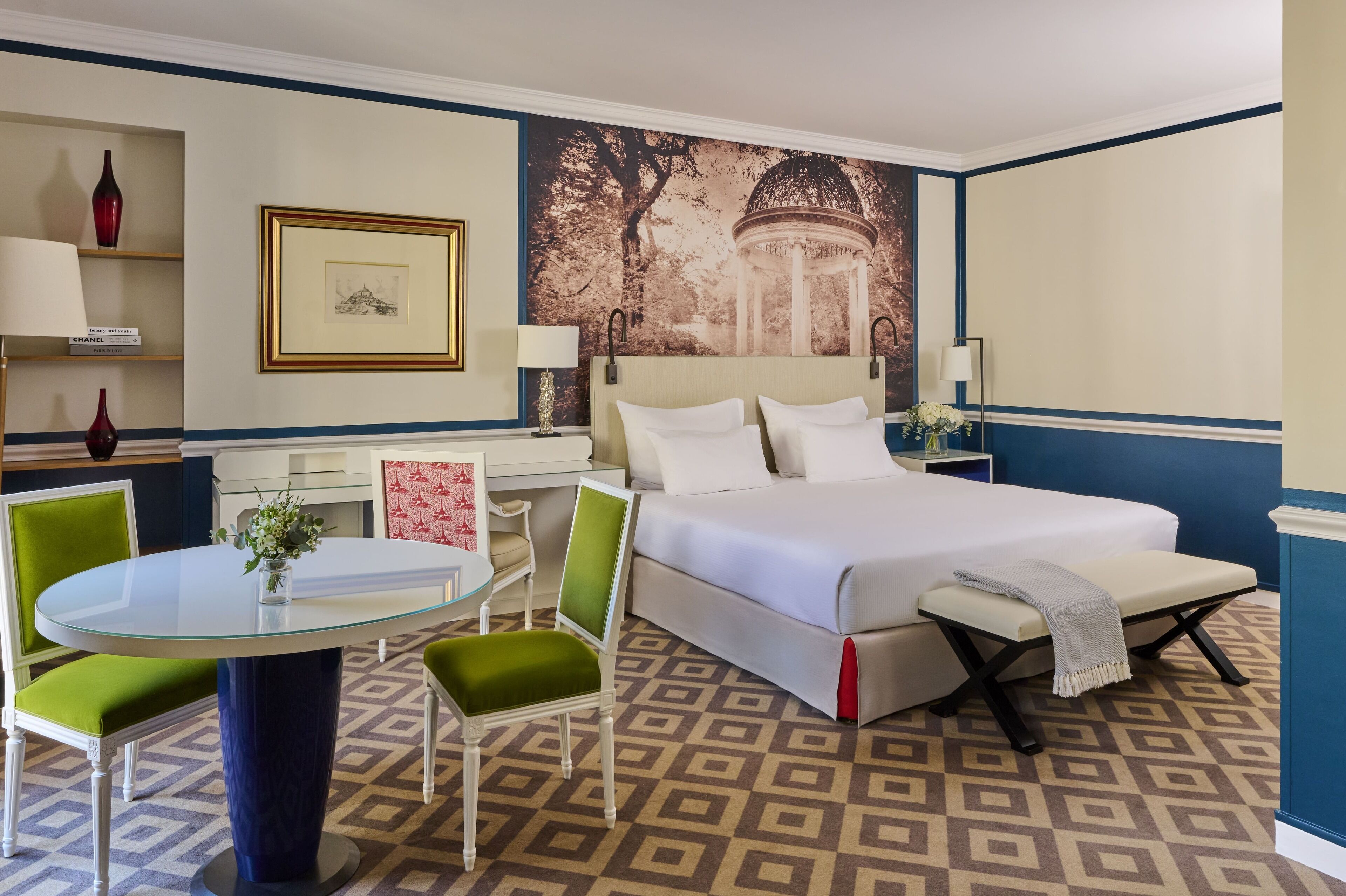 Quarto de hotel com uma cama, uma mesa redonda de vidro, duas cadeiras verdes, um sofá branco, uma pequena mesa com um vaso e uma grande tapeçaria na parede.