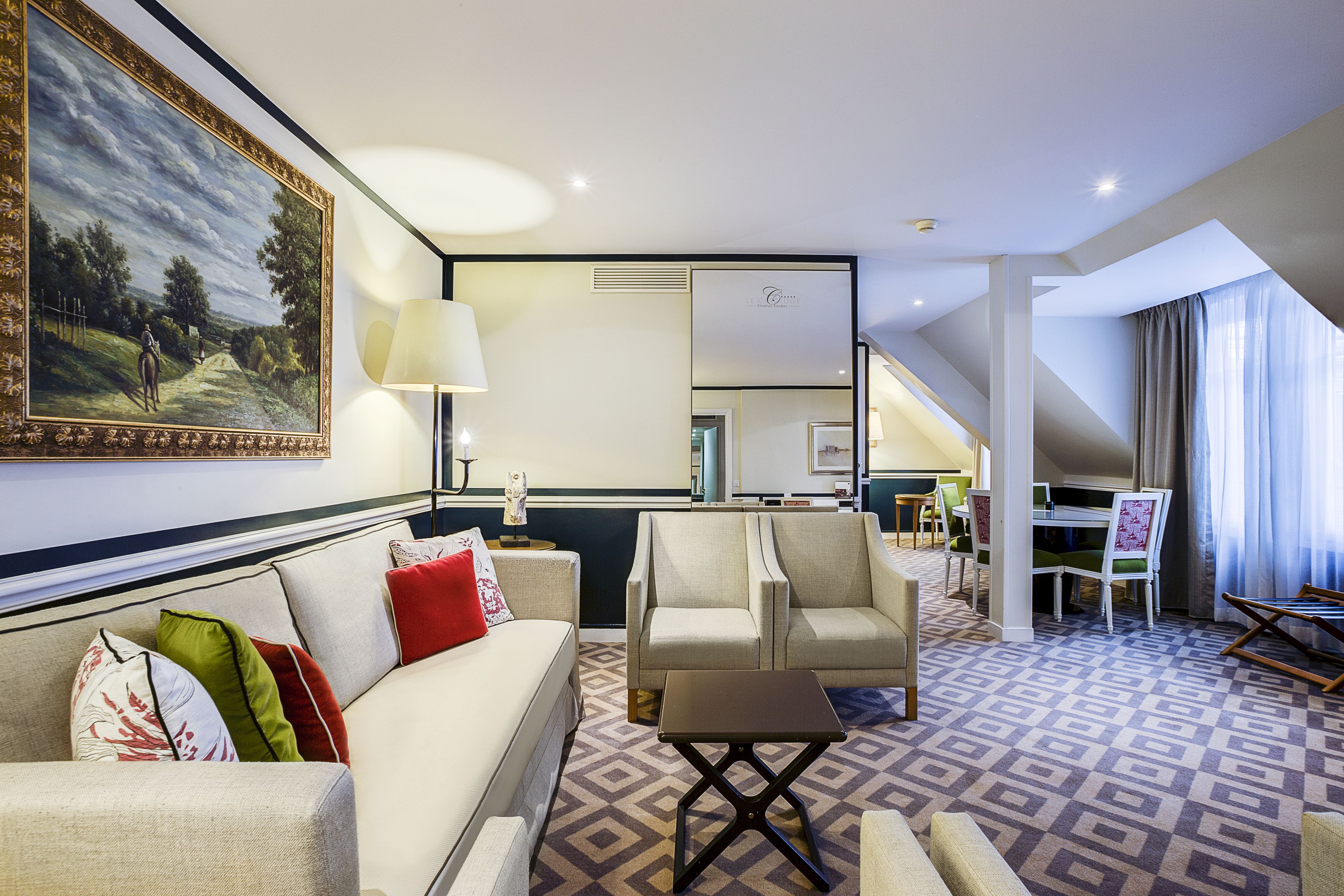 A hotel room with a sofa, armchairs, a small table, a painting, a lamp, and a view of the outdoors.