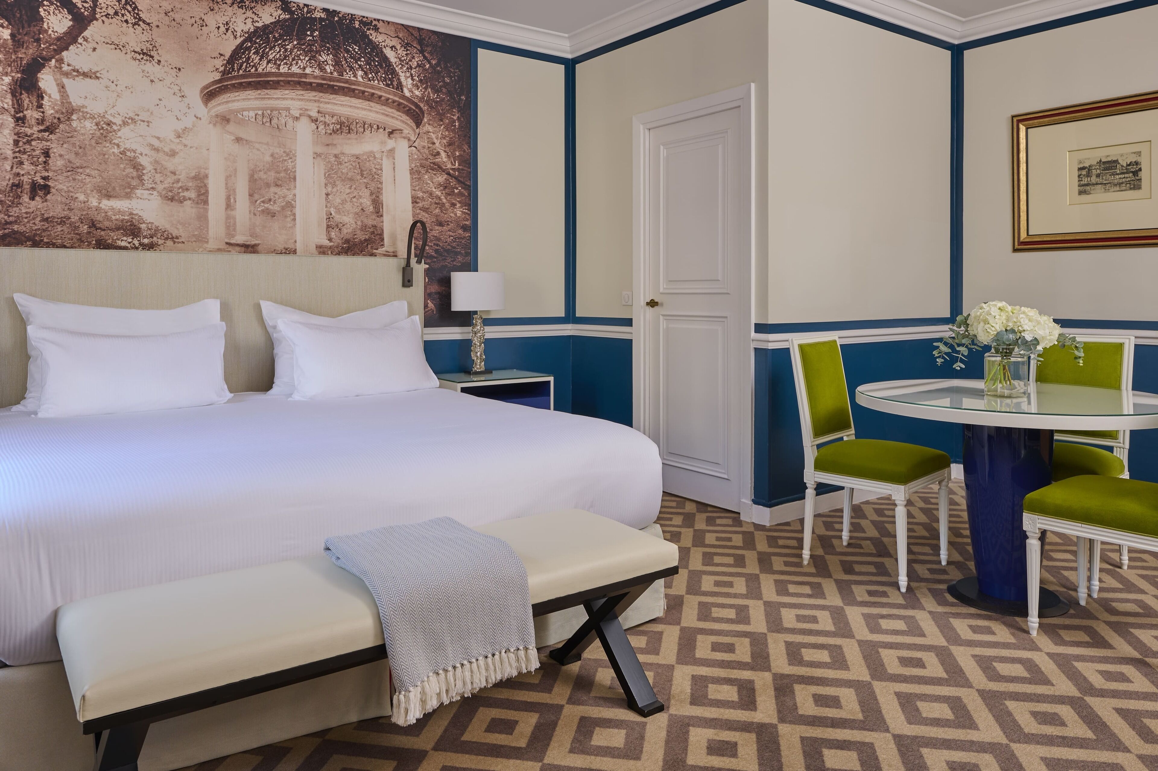 Habitación de hotel con una cama grande, un banco, una mesa redonda con sillas y una lámpara de pared.