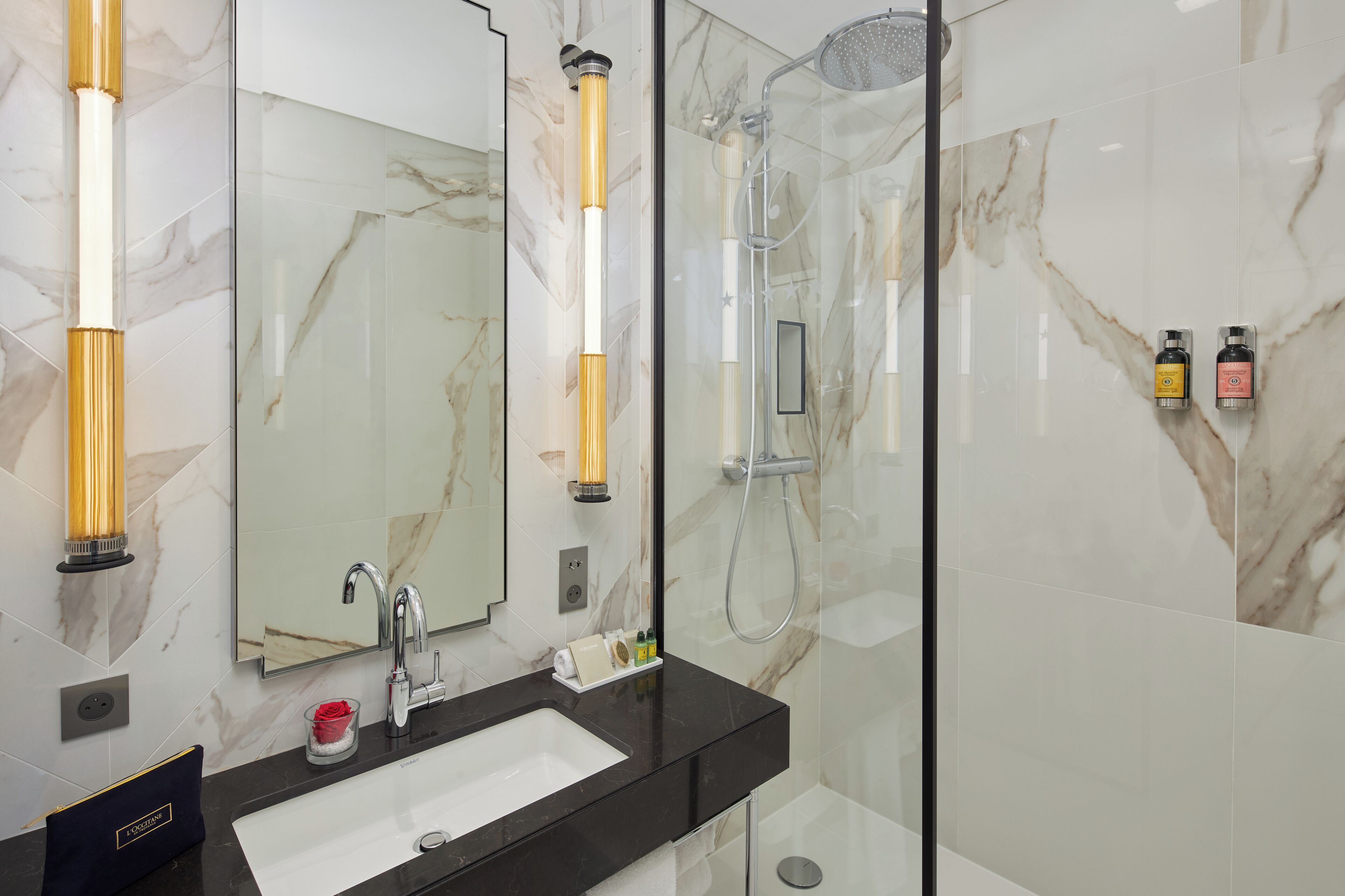 Un baño moderno con pared de azulejos de mármol, encimera negra, lavabo blanco, espejo grande y zona de ducha con mampara de vidrio.