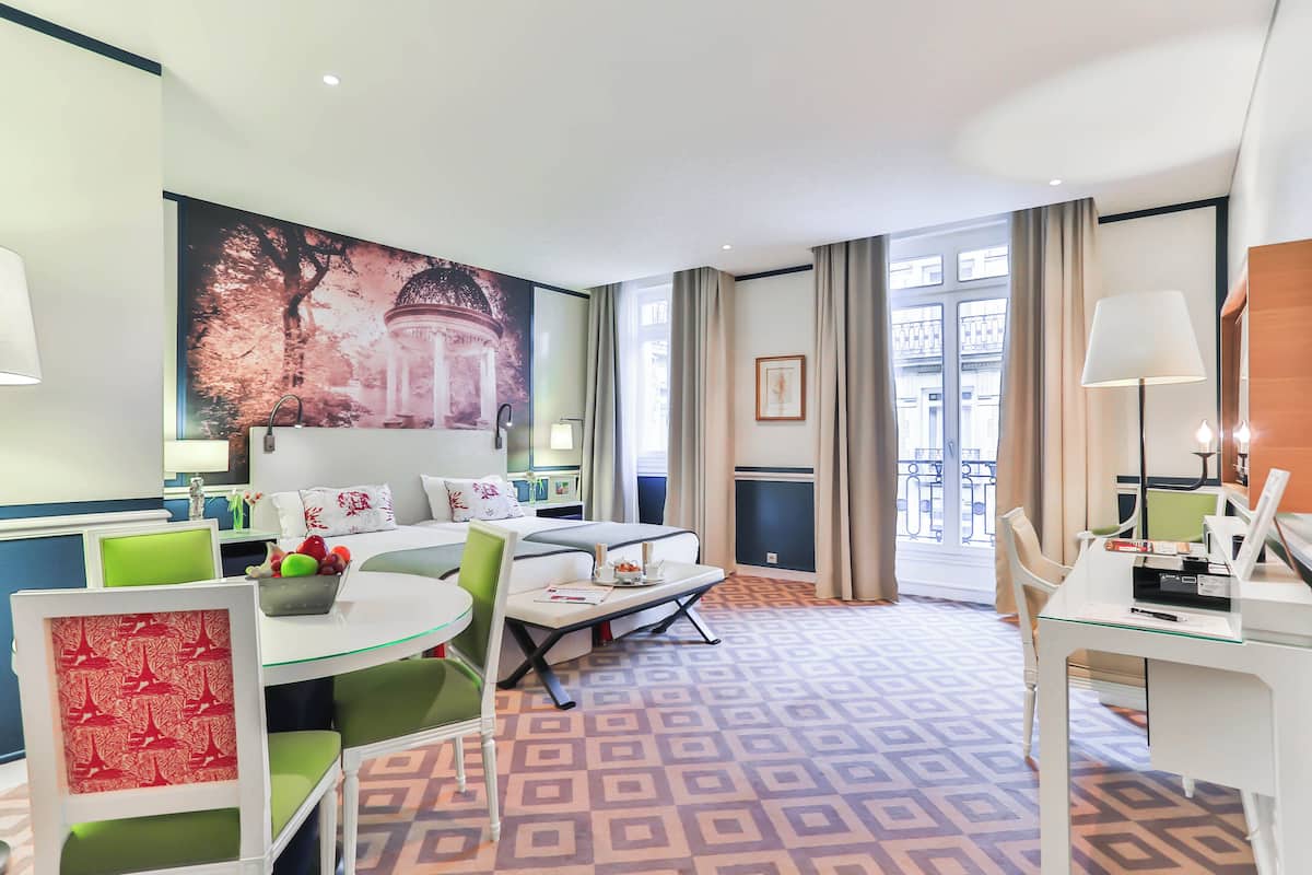 A hotel room with a bed, a dining area, a desk, and a large window.
