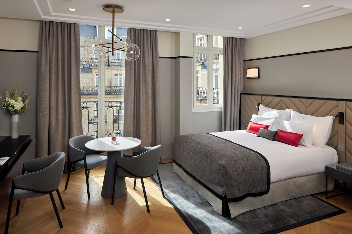 A modern hotel room with a large bed, a dining table with chairs, a view of the city through large windows, and a balcony with a railing.
