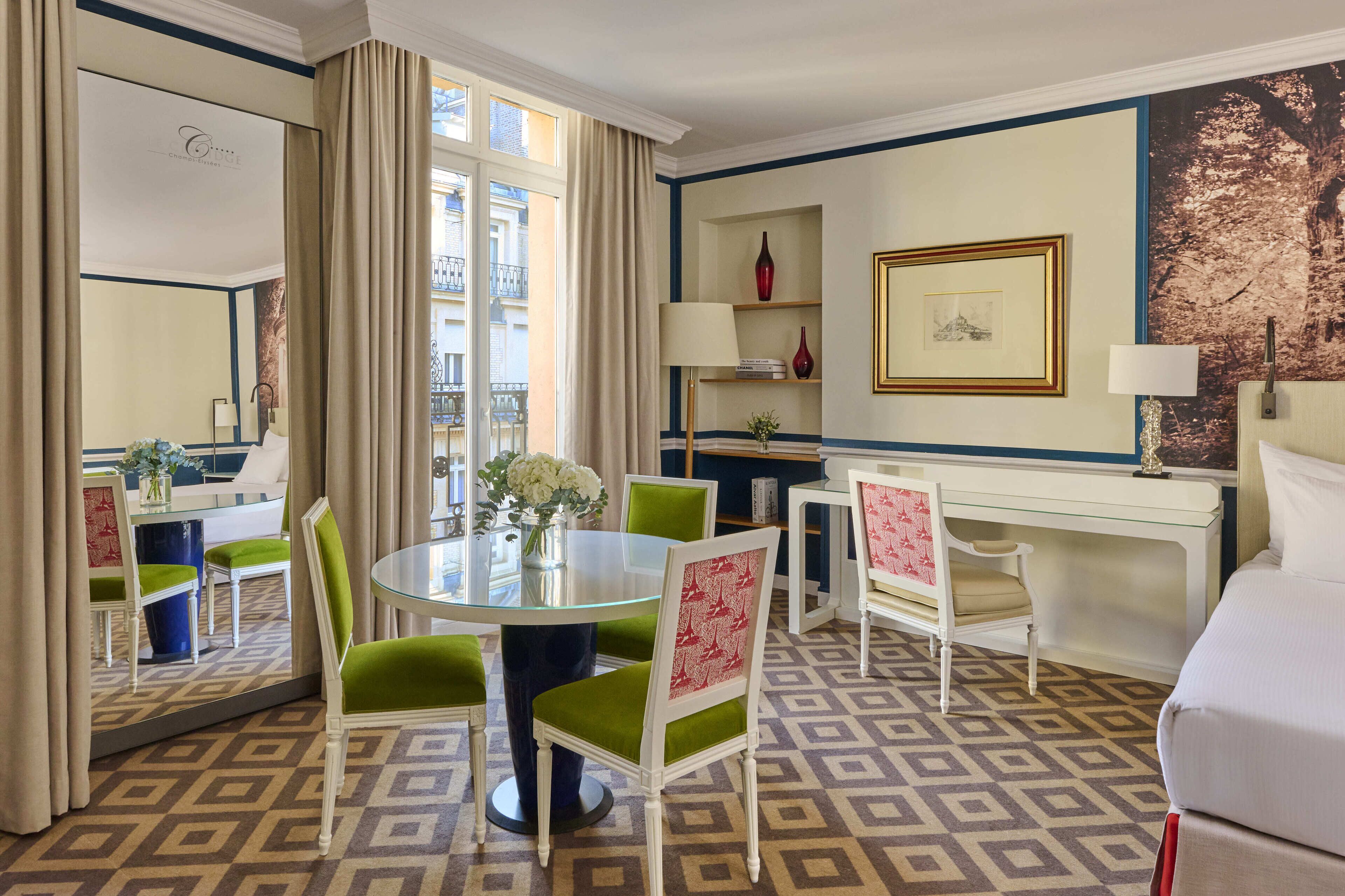 Habitación de hotel con una mesa redonda de vidrio, sillas blancas y verdes, una cama, un escritorio con lámpara, una estantería y un ventanal con cortinas.