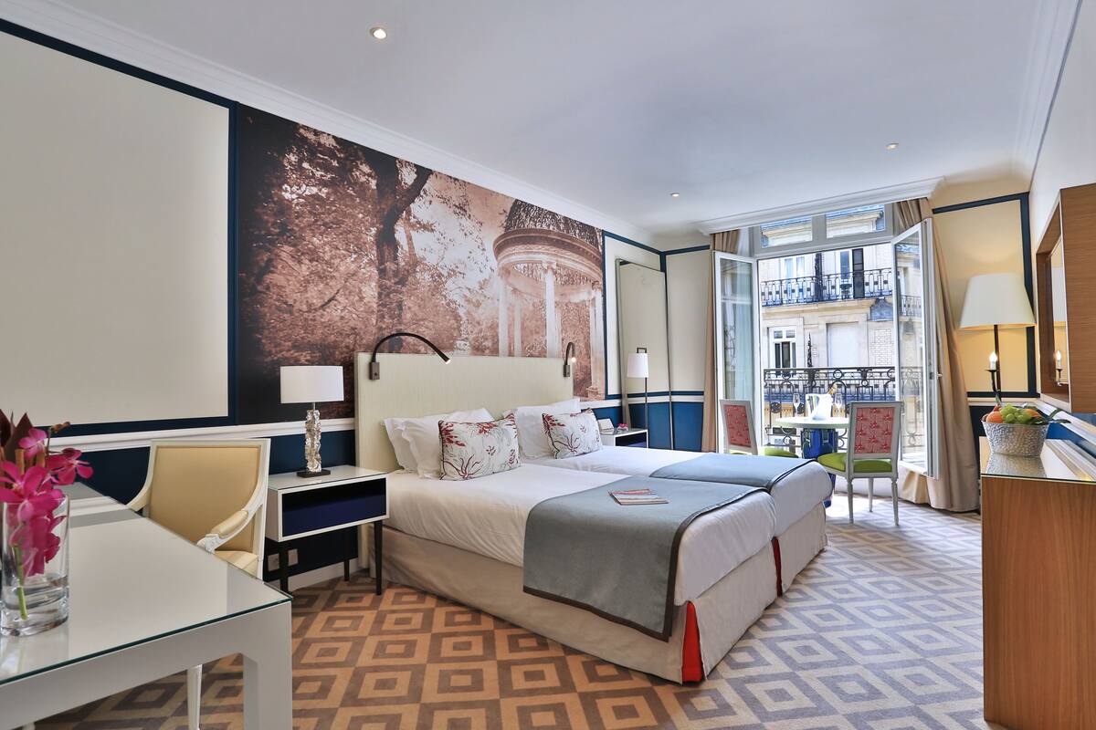 A hotel room with a large bed, a desk with a lamp, a chair, and a view of the outside balcony.