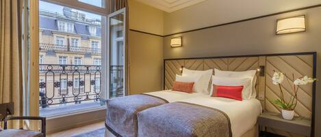 Hotelkamer met een groot bed, een klein nachtkastje, een stoel, een raam met uitzicht op gebouwen en een potplant.