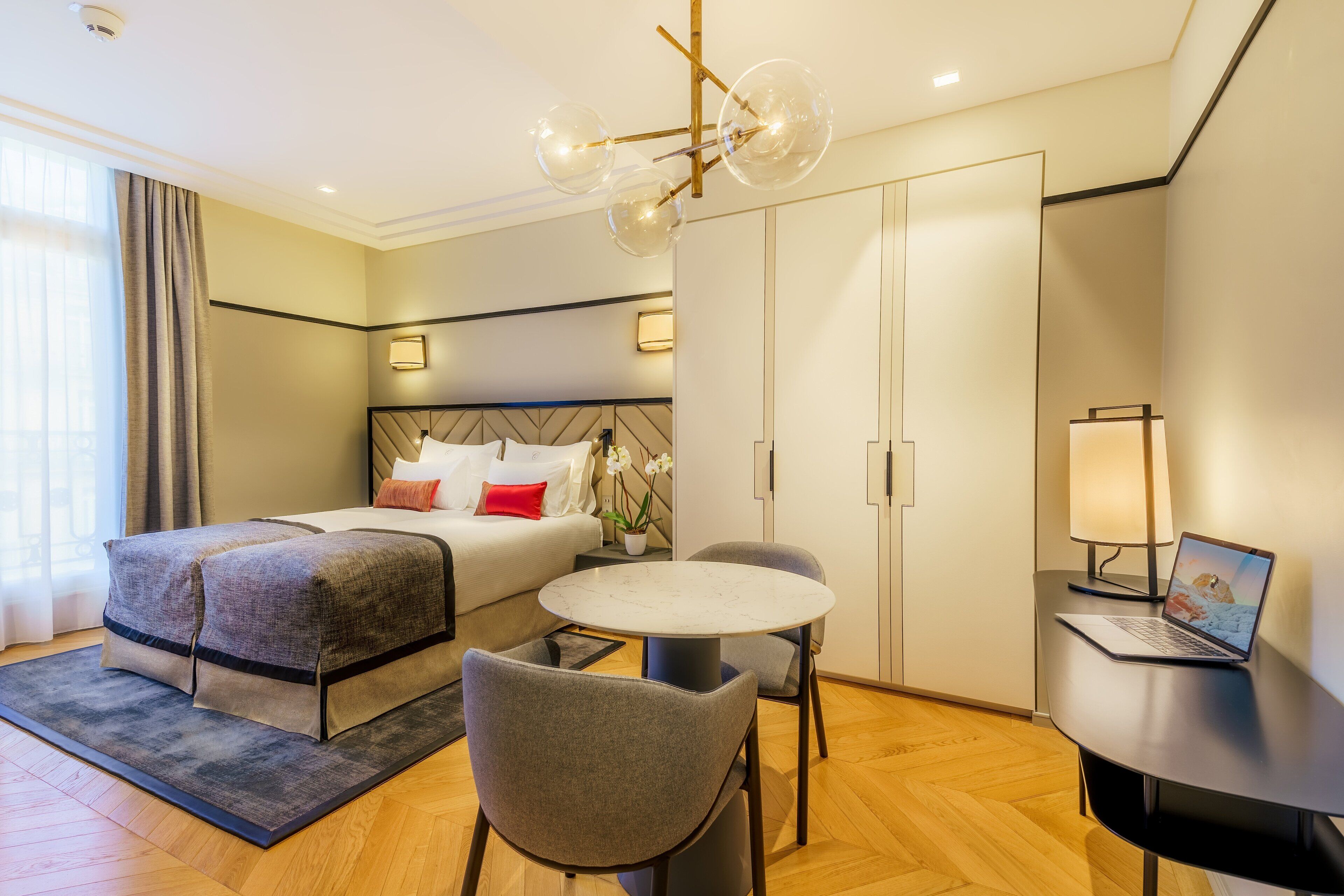 A modern hotel room with a bed, a round dining table, a laptop, and a desk lamp.