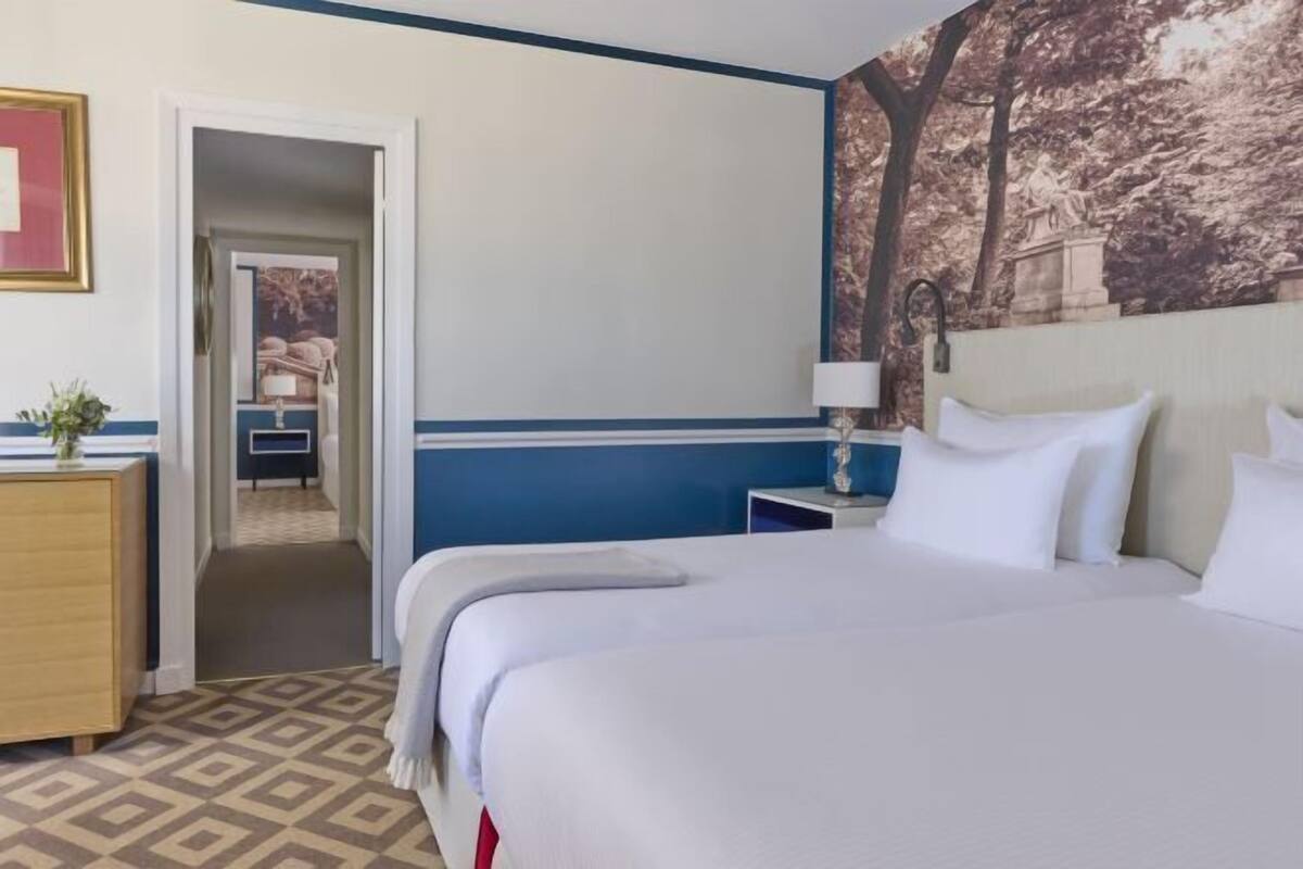 A hotel room with a bed, a nightstand, a lamp, a dresser, and a door leading to another room.