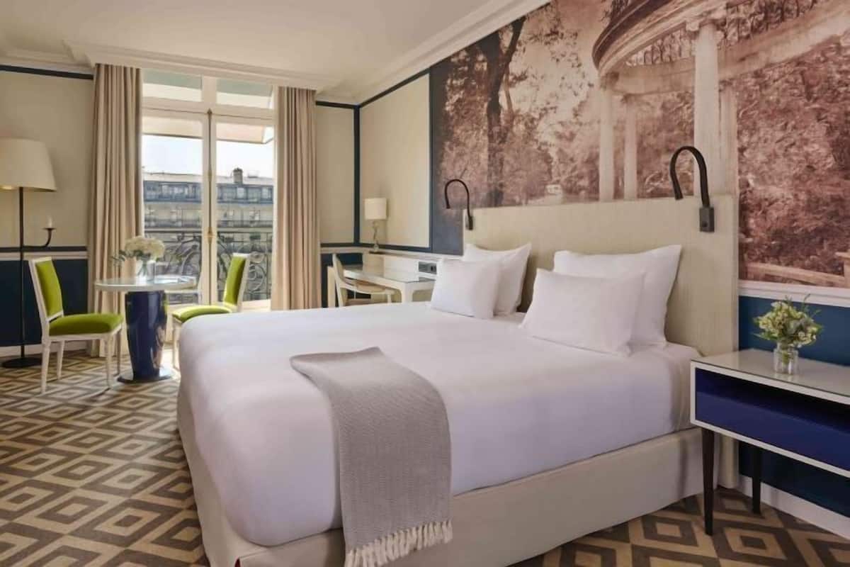 A hotel room with a large bed, a desk, a chair, a small table with a vase, and a view of the city through large windows.