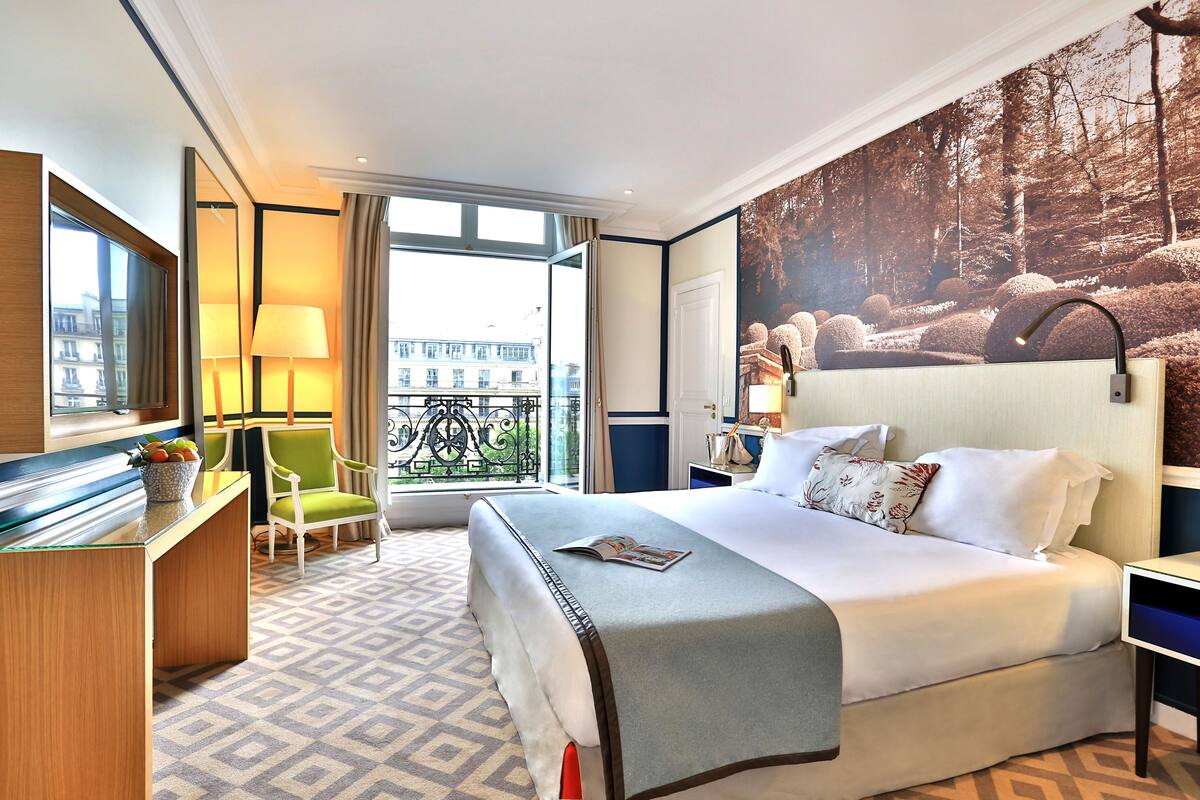 A hotel room with a large bed, a desk with a lamp, a chair, and a view of the city through the window.