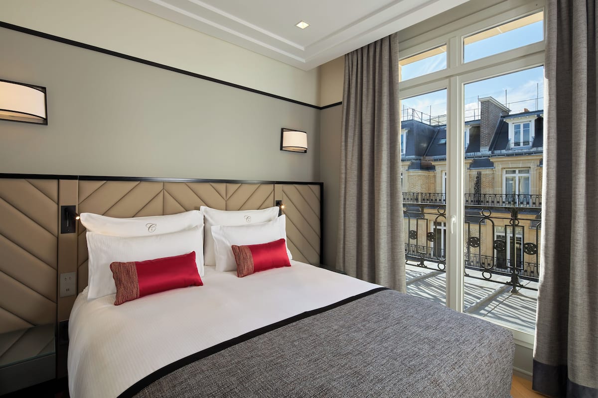A hotel room with a large bed, two bedside lamps, a view of the city through large windows, and a balcony with a railing.