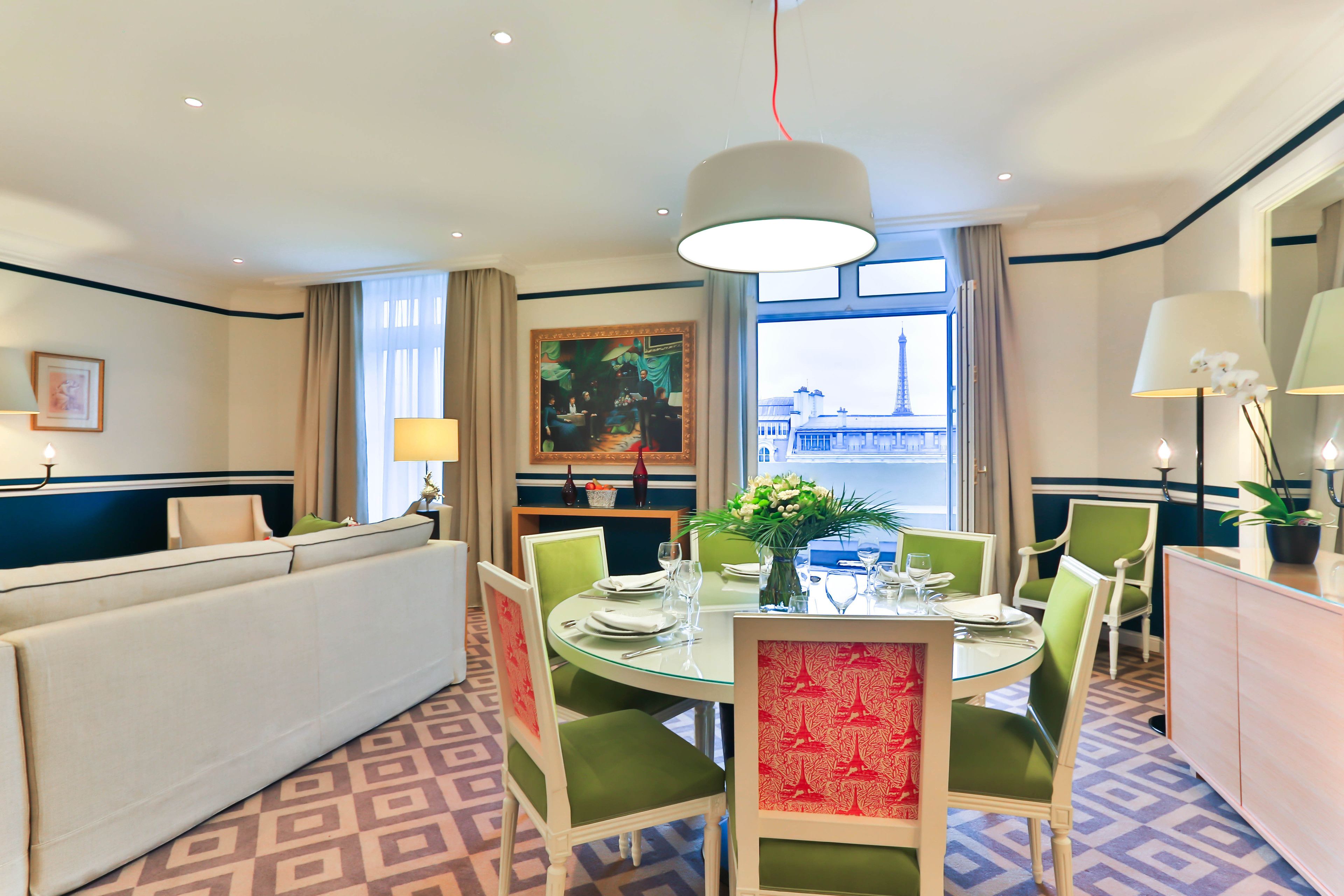 A hotel dining area with a round table set for four, green upholstered chairs, a glass-top table, and a large window with a view of a tower.