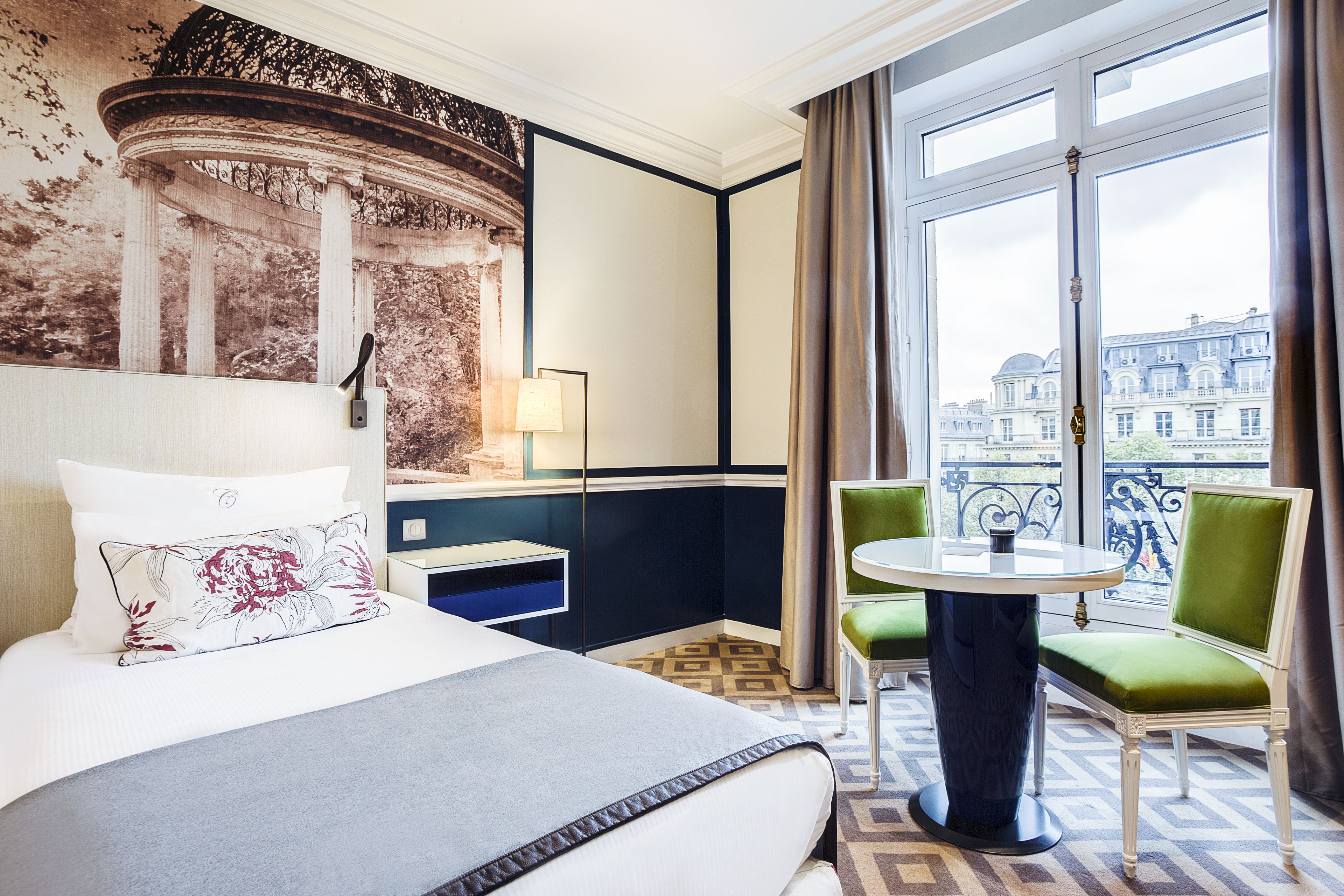 Habitación de hotel con cama, escritorio, silla, mesa y balcón con vistas a edificios.