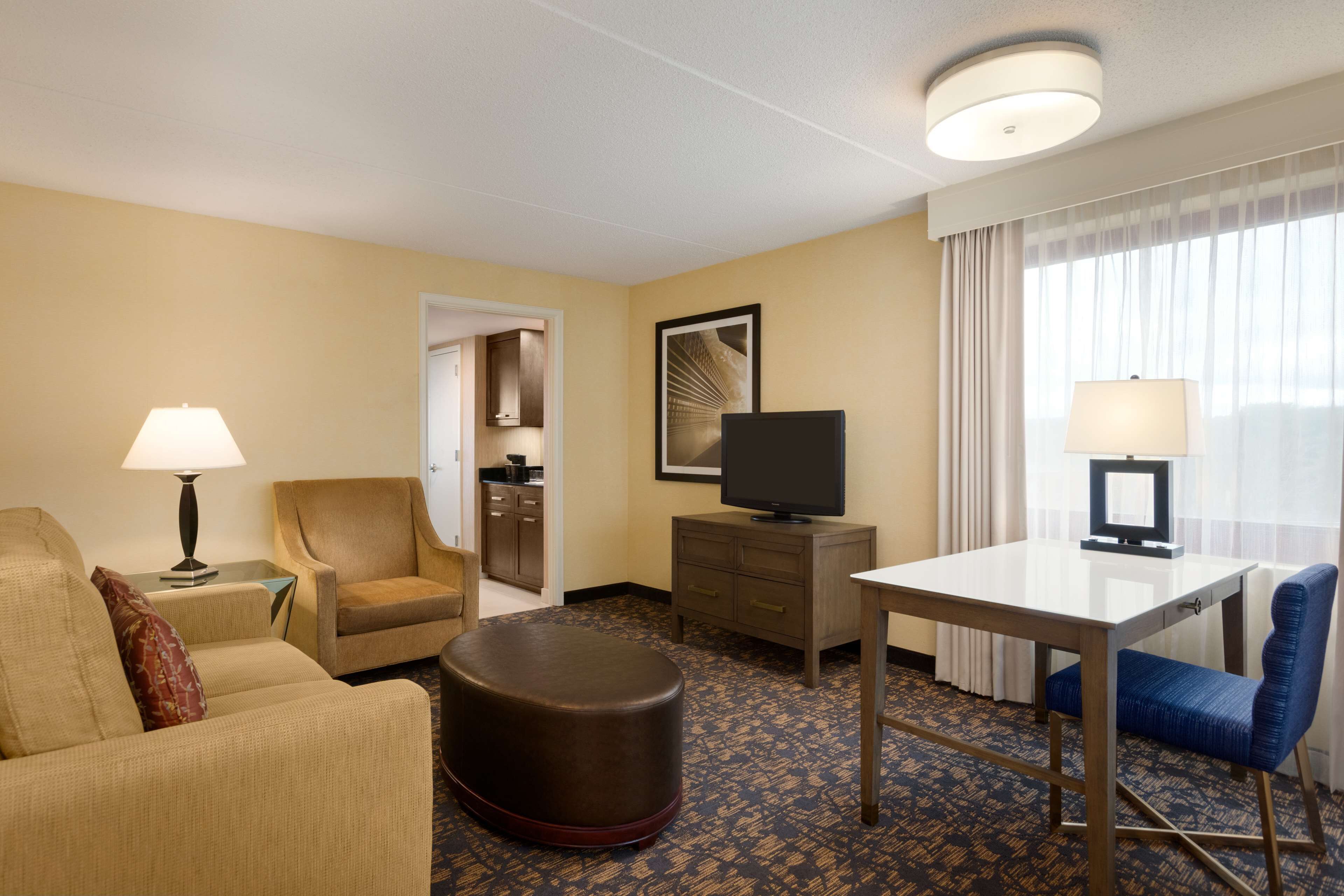 Habitació d'hotel amb sofà, butaca, otomana, televisió, escriptori i una finestra gran amb cortines.