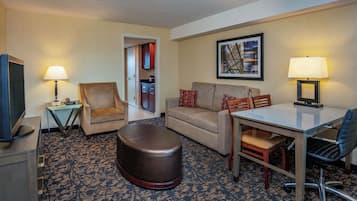 A hotel room with a sofa, ottoman, dining table, chairs, a TV, lamps, and a painting on the wall.