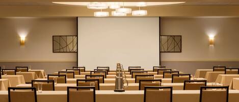 A conference room with rows of tables and chairs, a projector screen, and wall-mounted lighting.