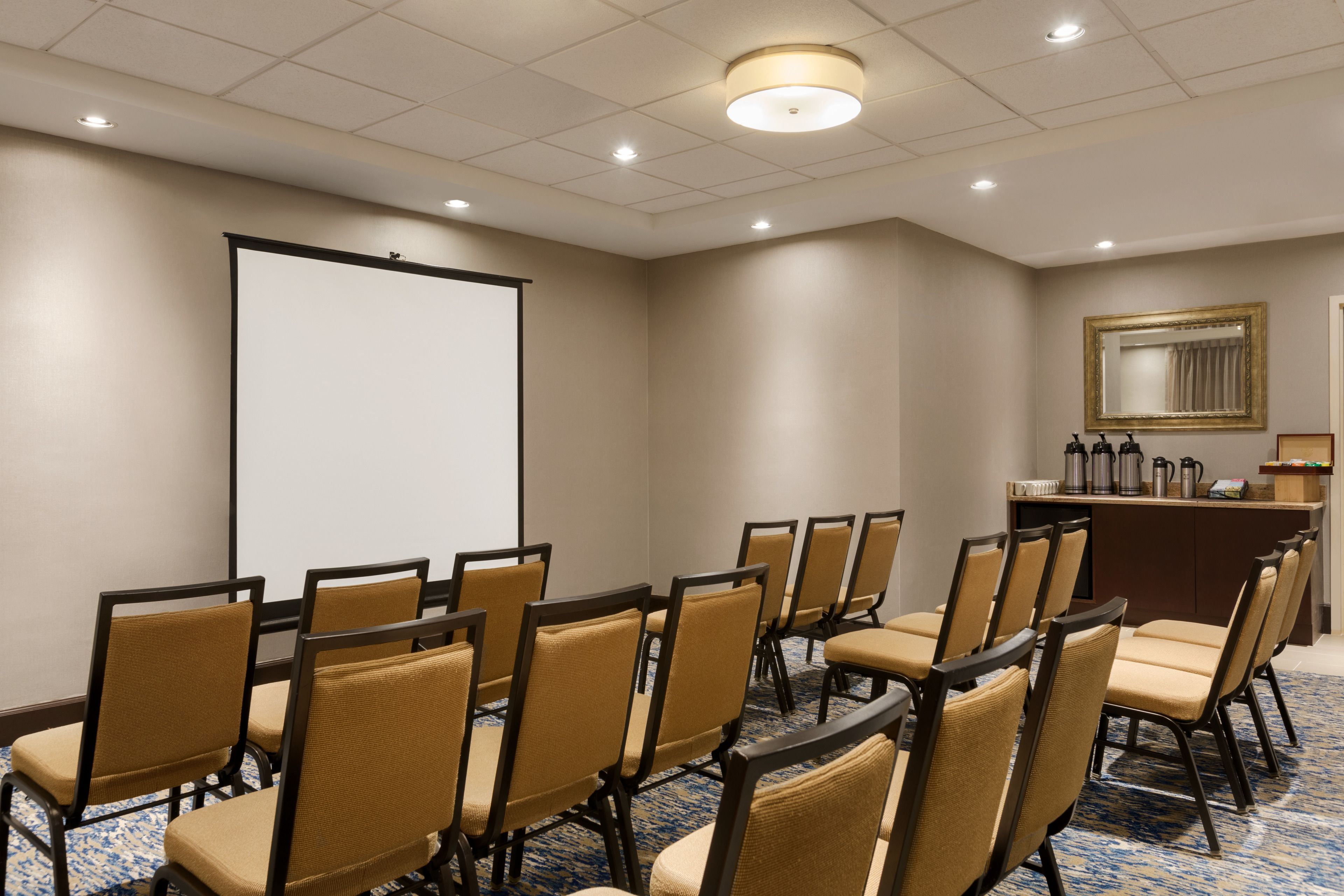 A conference room with rows of chairs, a projection screen, a coffee station, and a mirror on the wall.