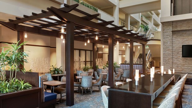 A spacious indoor area with a long dining table, multiple seating arrangements, and a ceiling with hanging lights. - Embassy Suites by Hilton Boston Waltham (Waltham)