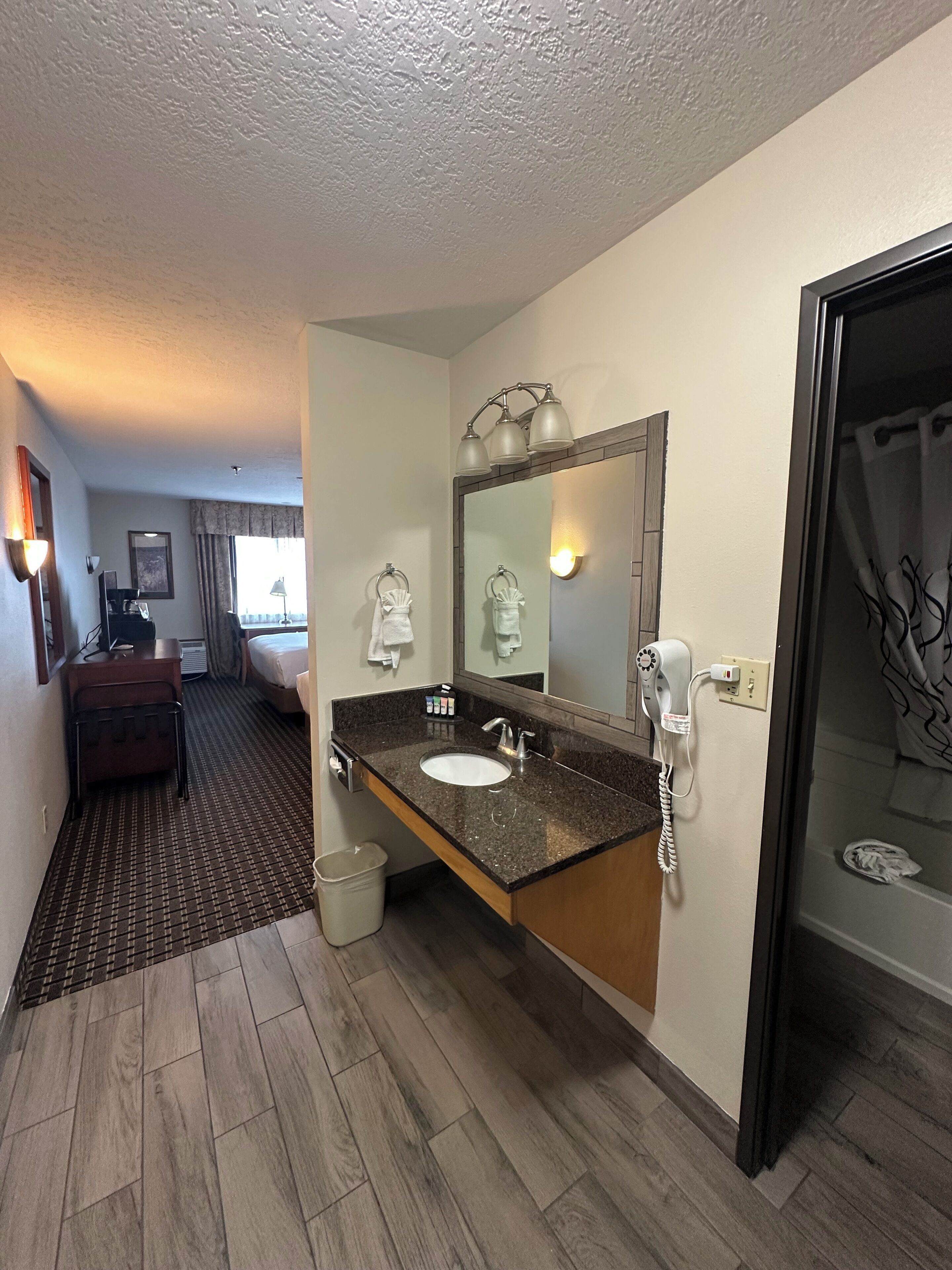 A hotel bathroom with a sink, mirror, and shower. There is a hairdryer on the wall and a towel rack. The floor is wooden, and there is a window with curtains.