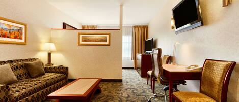 A hotel room with a sofa, a wooden coffee table, a desk with a chair, a television mounted on the wall, and framed artwork on the walls.