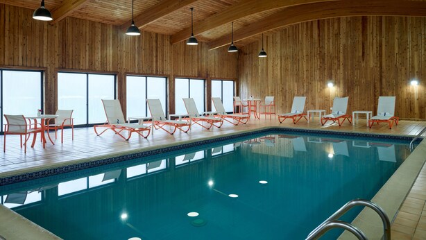 An indoor swimming pool with wooden walls, multiple lounge chairs, and ceiling lights.