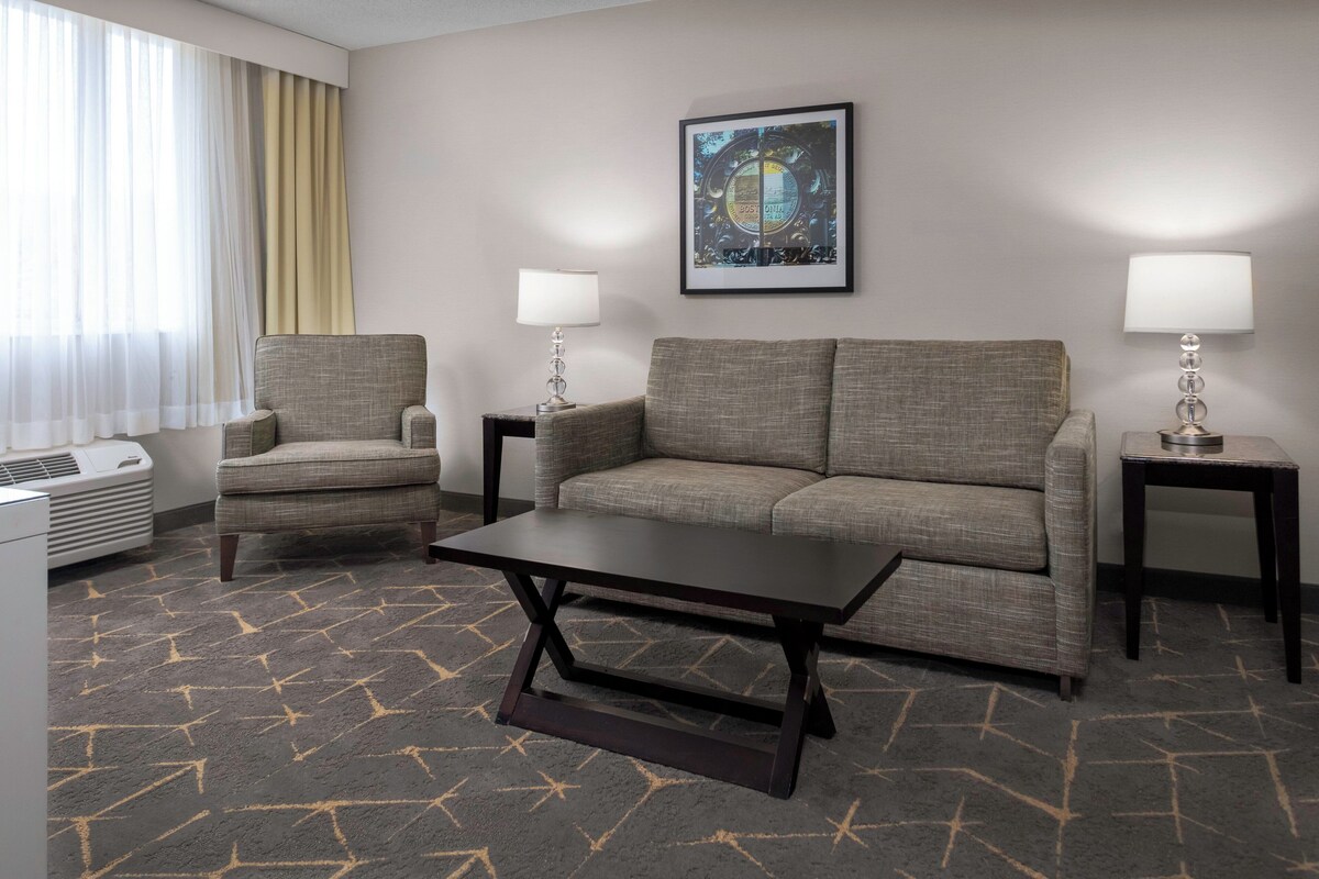 A hotel room with a sofa, armchair, and a coffee table. There is a picture on the wall and a lamp on each side of the sofa.