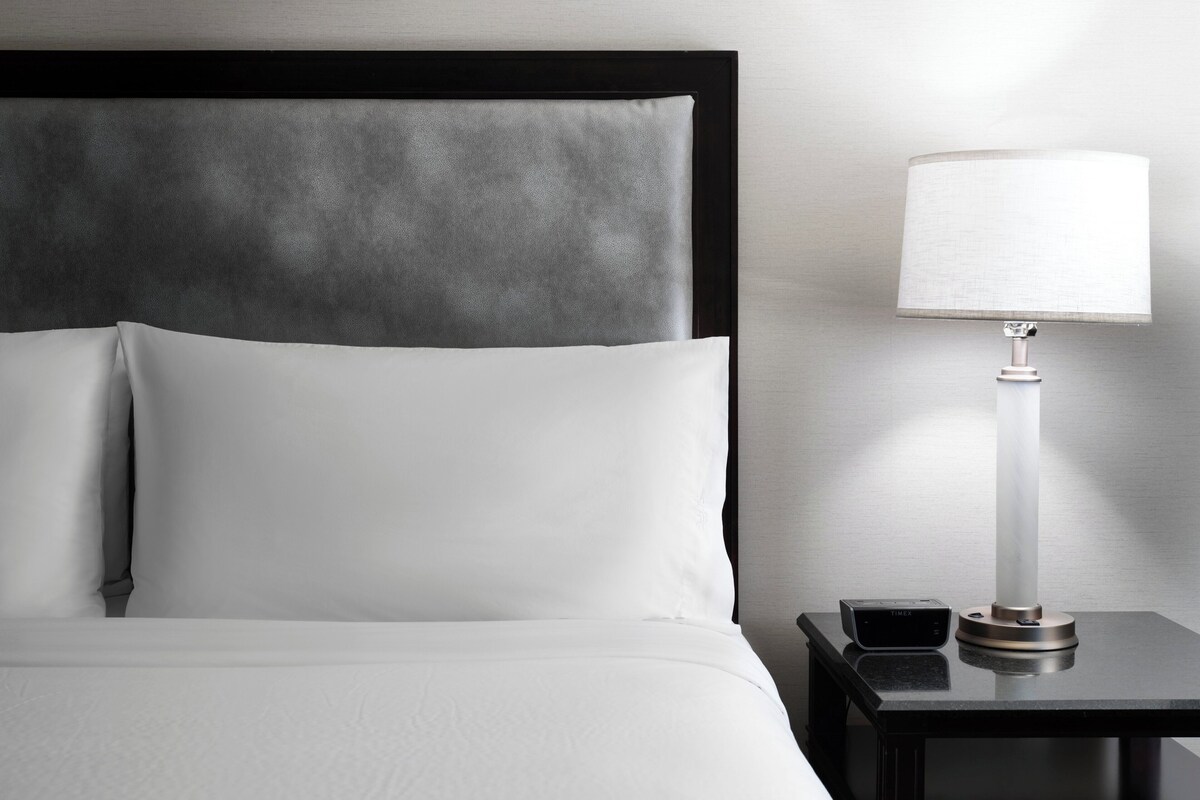 A neatly made bed with white linens, a black headboard, and a bedside table with a lamp and a small device.
