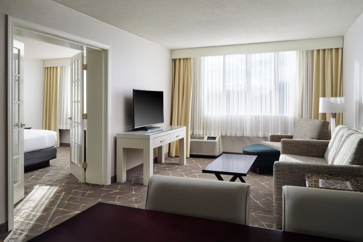A hotel room with a bed, a desk with a television, a seating area with a sofa and armchairs, and a large window with curtains.