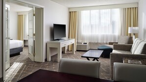 A hotel room with a bed, a desk with a television, a seating area with a sofa and armchairs, and a large window with curtains.