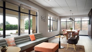 A modern living room with a grey sofa, orange cushion, and a wooden coffee table. There is a dining area with a table and chairs, and large windows offering a view of the outdoors.