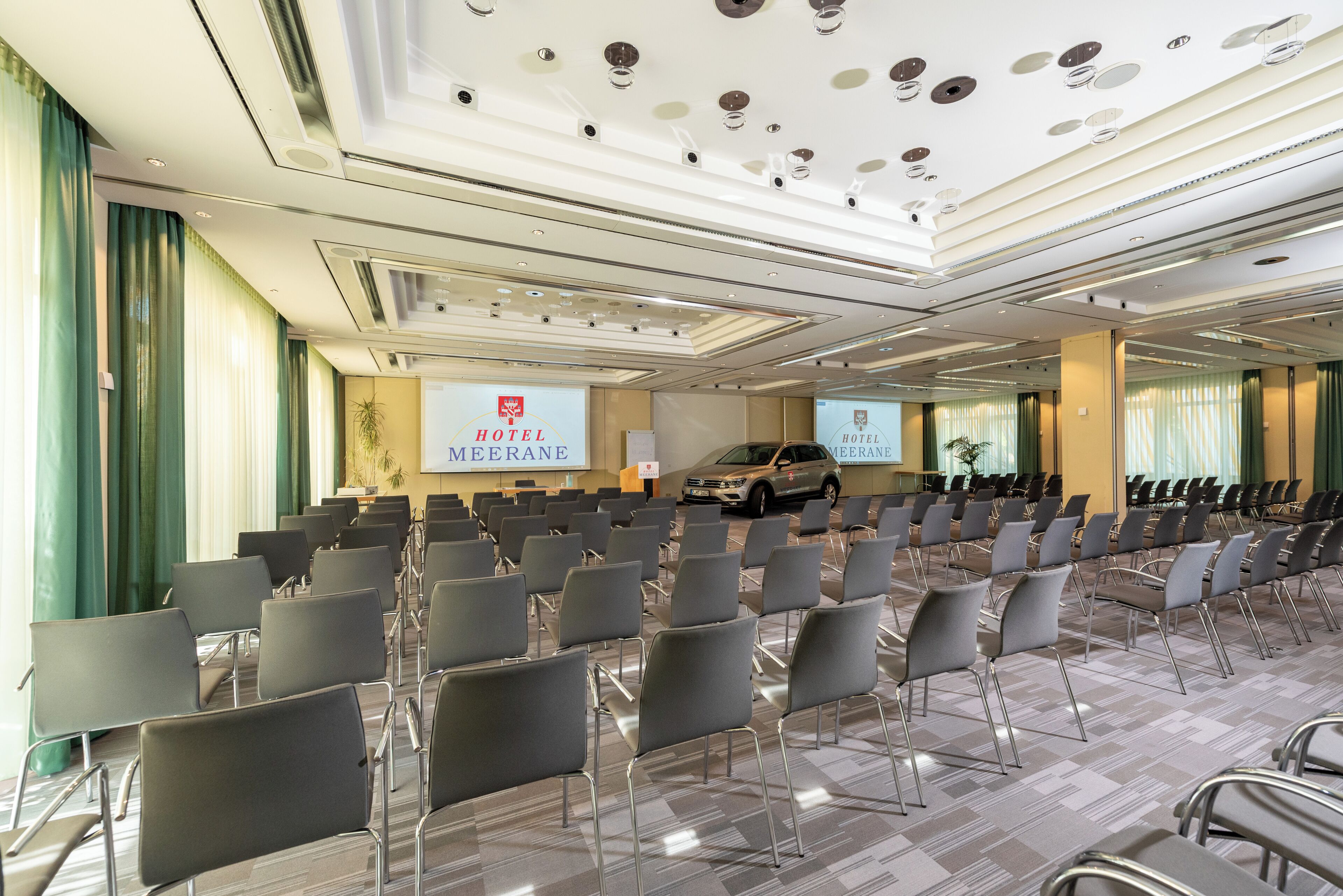 Une salle de conférence d’hôtel avec des rangées de chaises, un pupitre et une voiture garée à l’extérieur.