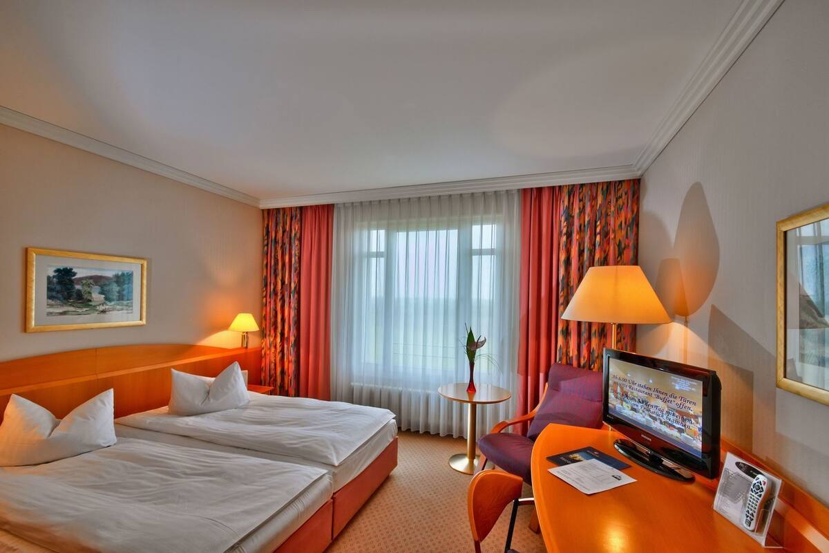 Habitación de hotel con dos camas, un escritorio con televisor, una mesita con un florero y una ventana con cortinas.