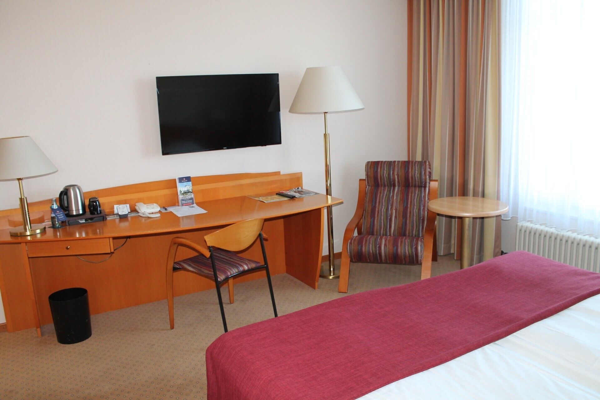 A hotel room with a bed, a desk with a chair, a television, a lamp, a striped armchair, and a small table.