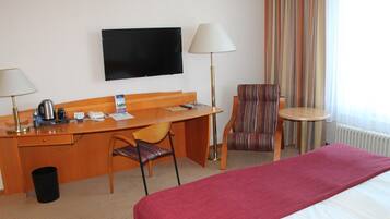 A hotel room with a bed, a desk with a chair, a television, a lamp, a striped armchair, and a small table.