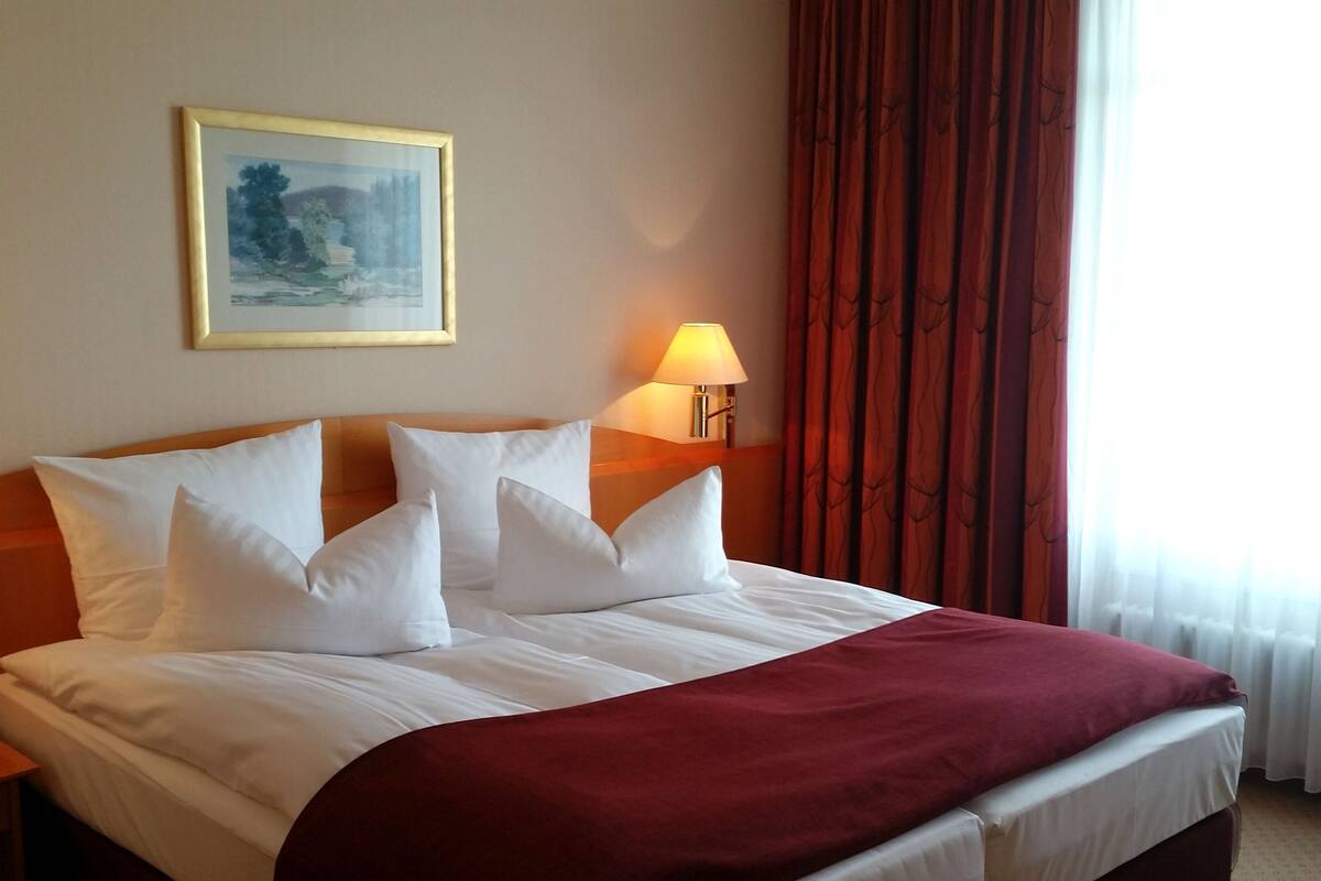 Una cama bien hecha con sábanas blancas y un plaid rojo, una lámpara de noche y un cuadro en la pared.