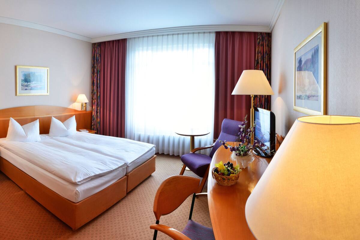 Habitación de hotel con una cama grande, un escritorio con lámpara, una silla y una pequeña mesa con una cesta de frutas.