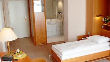 Habitación de hotel con cama, escritorio con lámpara, teléfono, silla y baño visible a través de una puerta abierta.