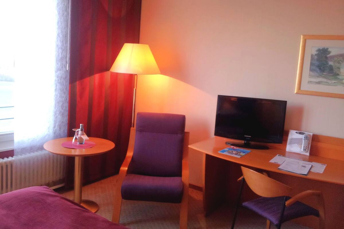 Una habitación de hotel con una cama, un escritorio con computadora, una silla, una lámpara y una ventana con cortinas.