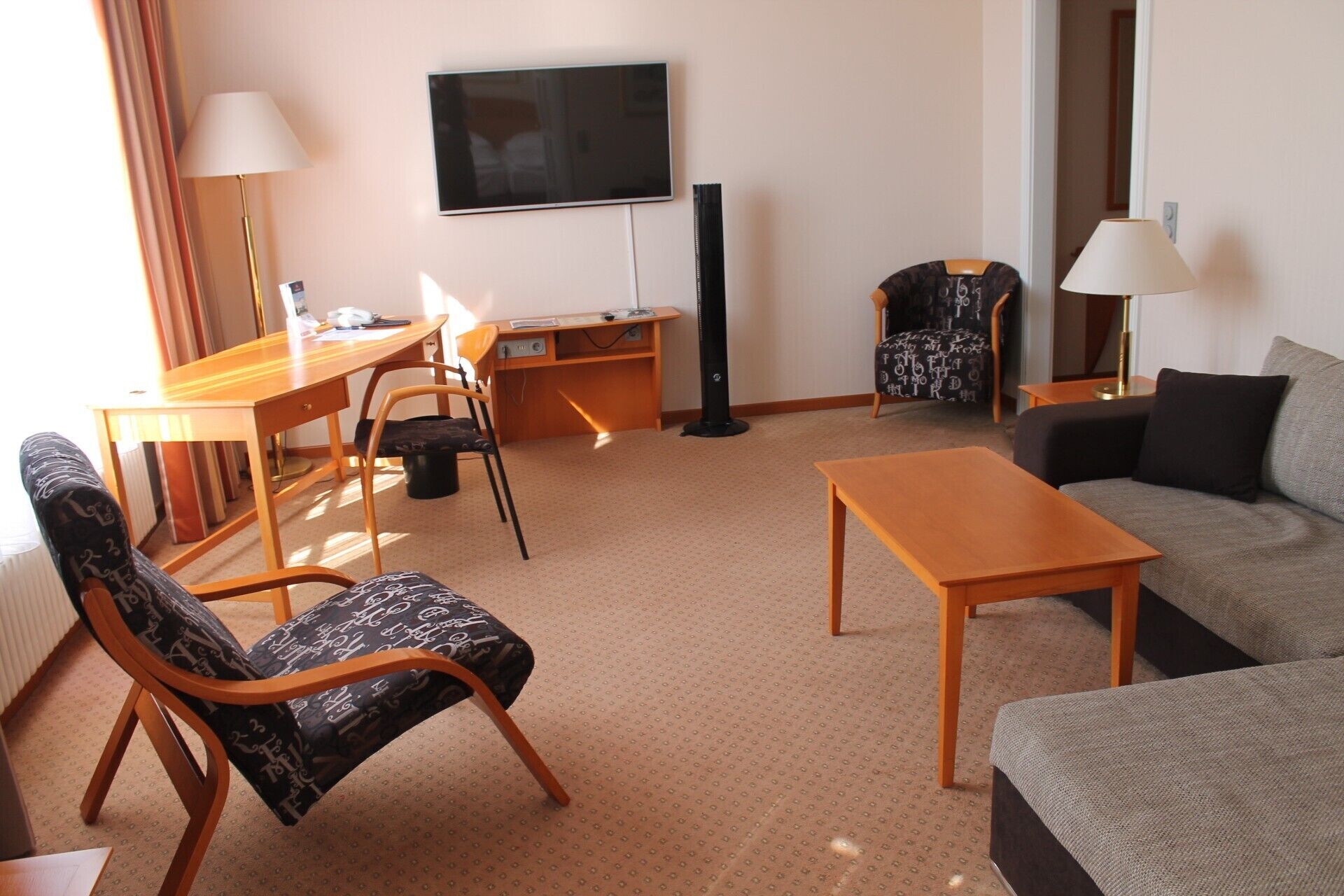 Een hotelkamer met een bank, een bureau, een stoel, een televisie en een ventilator.