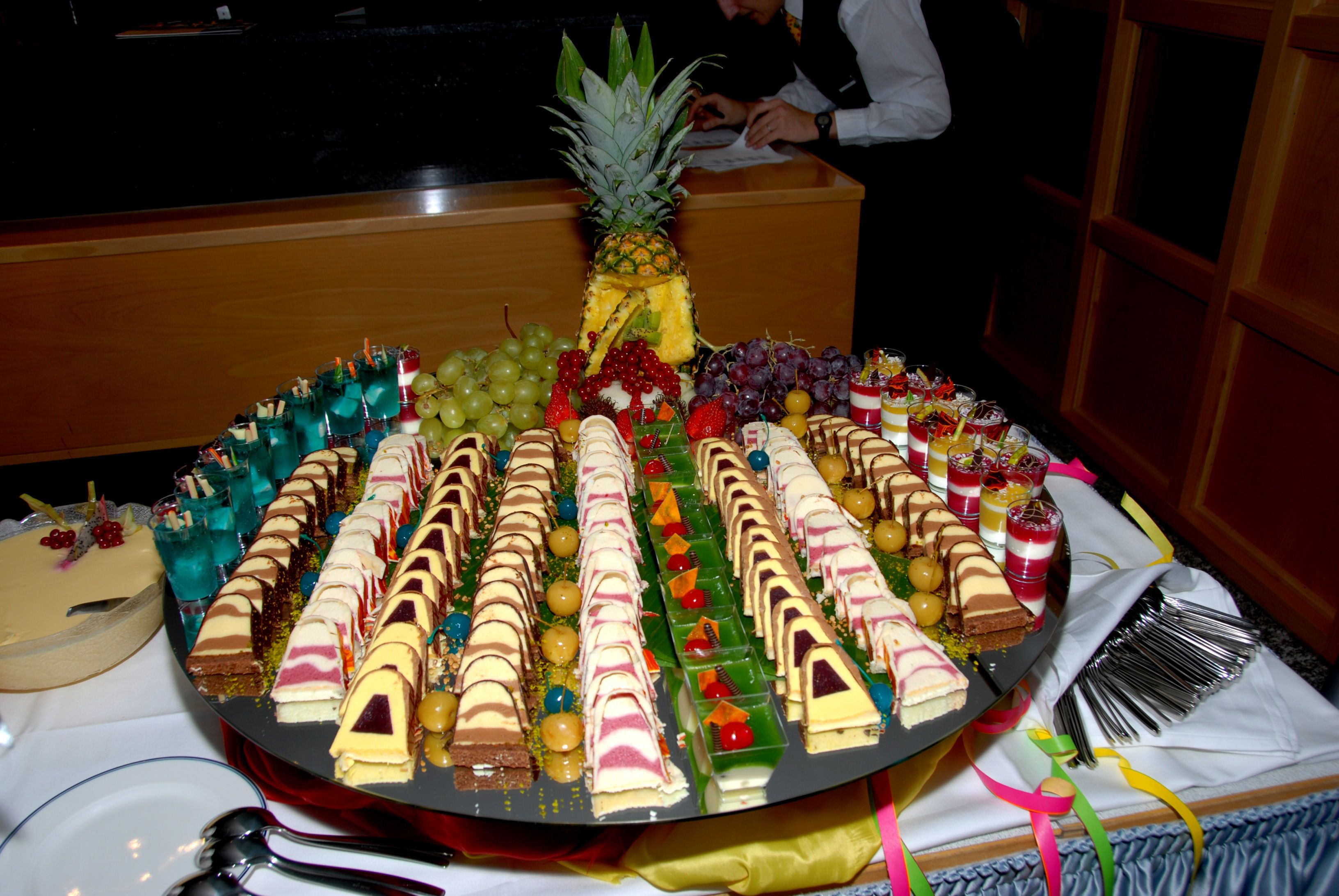 A large, round dessert platter with various types of cakes and pastries, a pineapple, and an assortment of fruits and beverages.