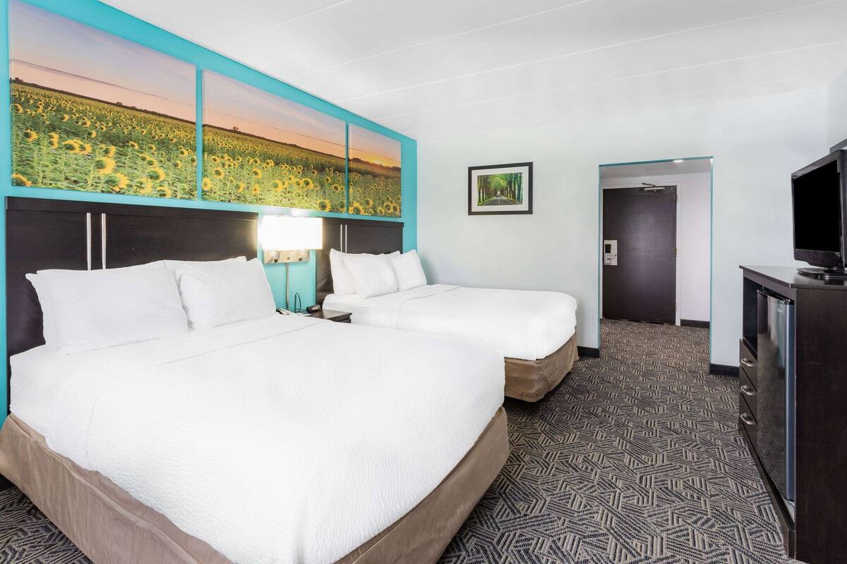 A hotel room with two beds, a TV, and a painting of a sunflower field.